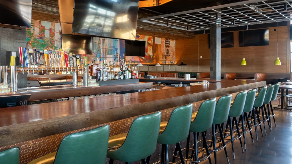 Modern bar interior with long wooden counter, green stools, and liquor display.