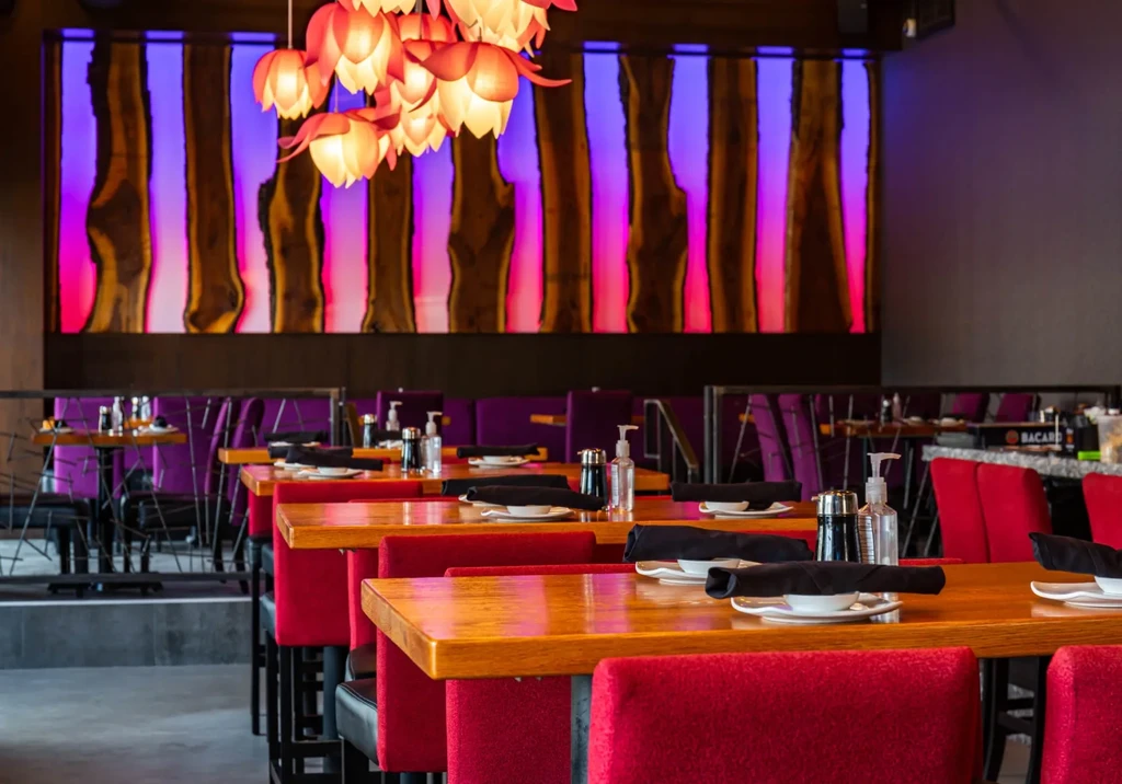 Modern restaurant with red chairs, purple booths, backlit wood wall, flower lights.