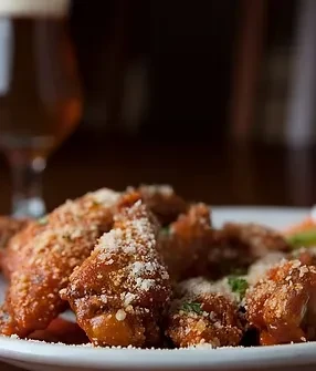 Close-up of chicken wings, reddish sauce, Parmesan, herbs, blurred beer.