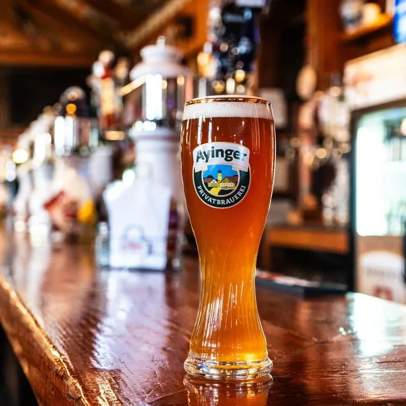 Pilsner glass of amber Ayinger beer with frothy head at a bar.