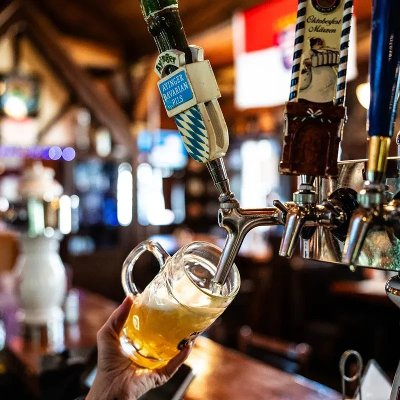 Hand filling a glass stein with Ayinger Bavarian Pils from a tap.