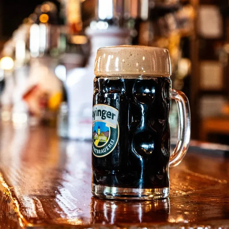 Ayinger dark beer mug with frothy head on a reflective wooden bar.