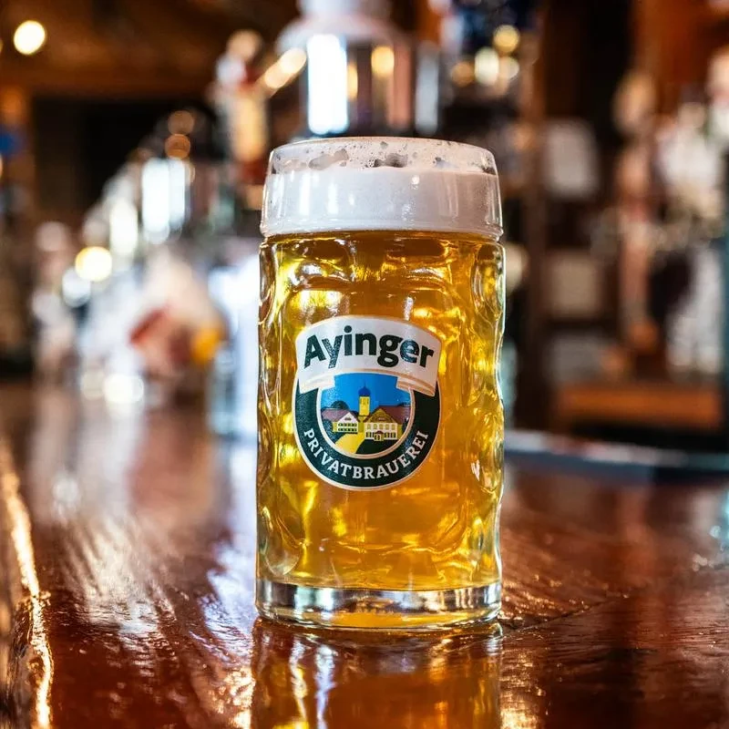Frothy Ayinger beer stein on a dark bar, blurred warm background.