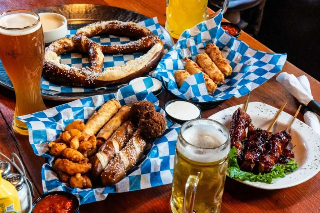 Pretzel, fried appetizers, meat skewers, and beer on a wooden table.