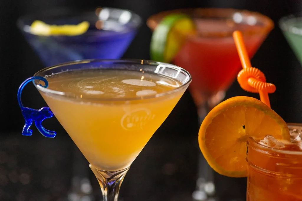 Colorful cocktails, one with a blue monkey clip, in a dark setting.