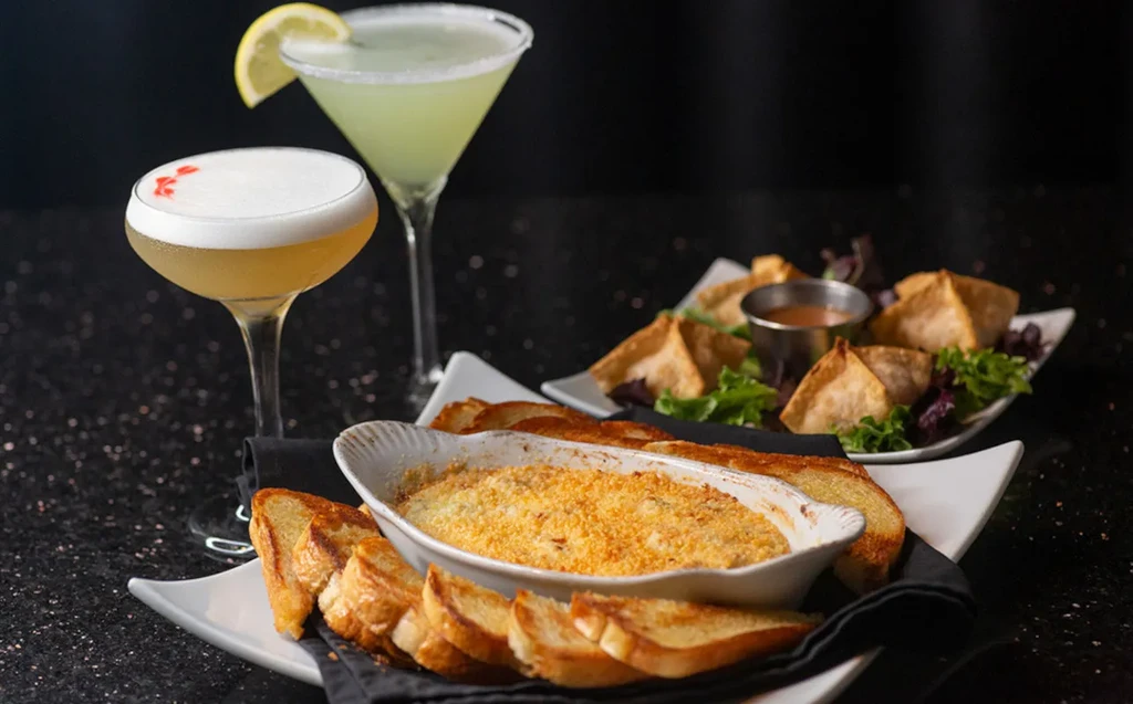 Baked dip, two cocktails, and fried pastries on a dark counter.