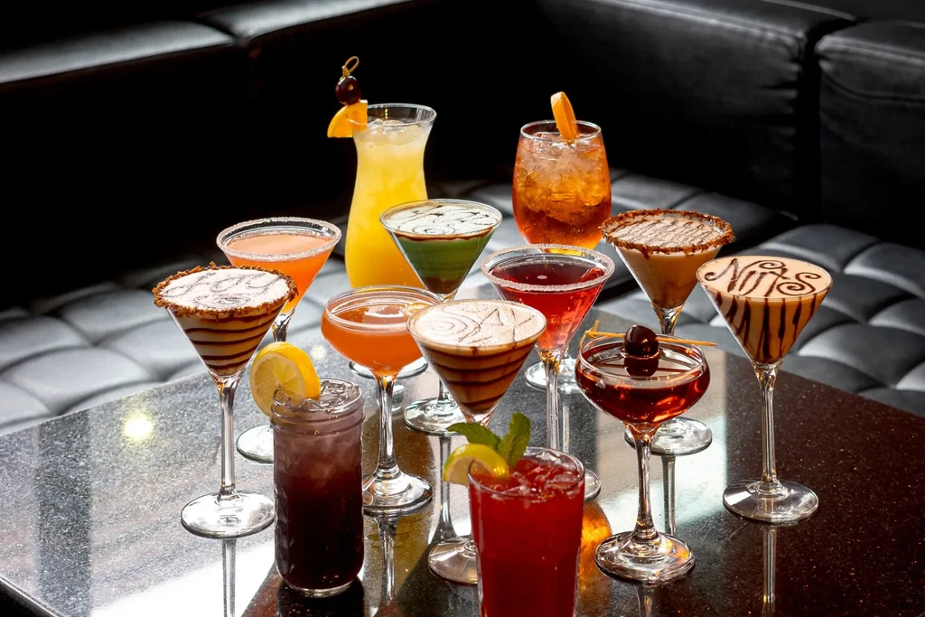 Ten diverse garnished cocktails with 'Nils' on one, on a dark table.