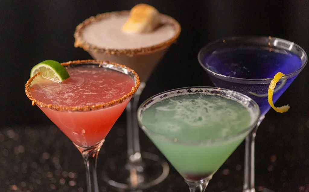 Four colorful cocktails in martini glasses with various garnishes.