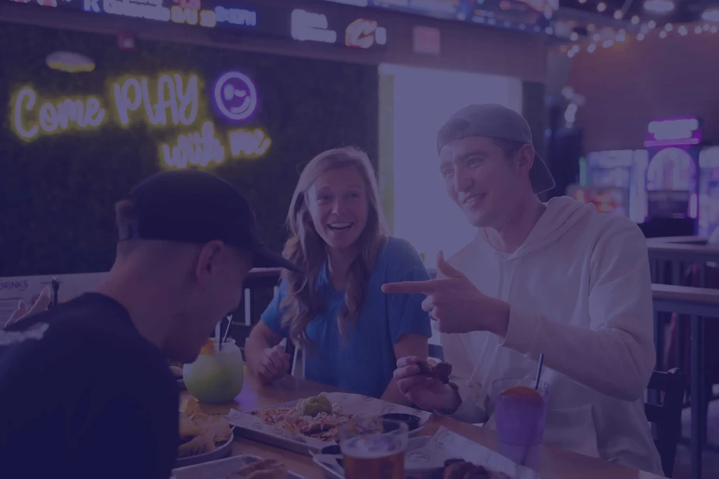 Three young adults laugh, point, and eat at a purple-lit bar.