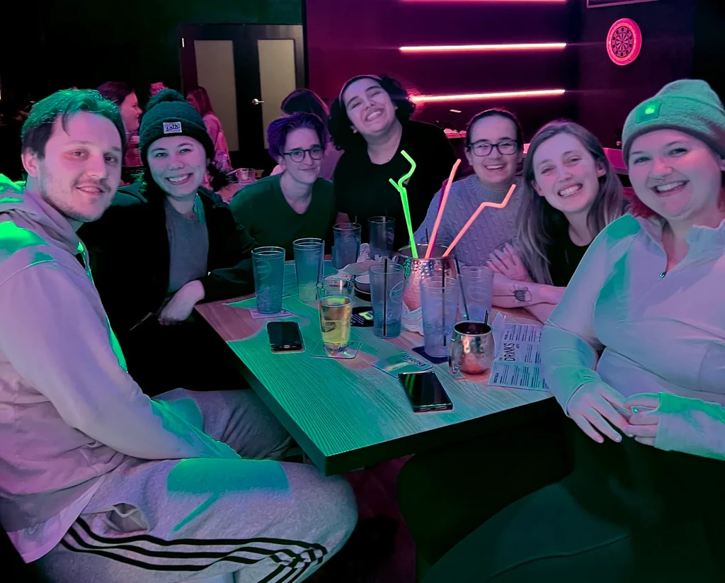 Seven smiling people with drinks in a neon-lit bar.