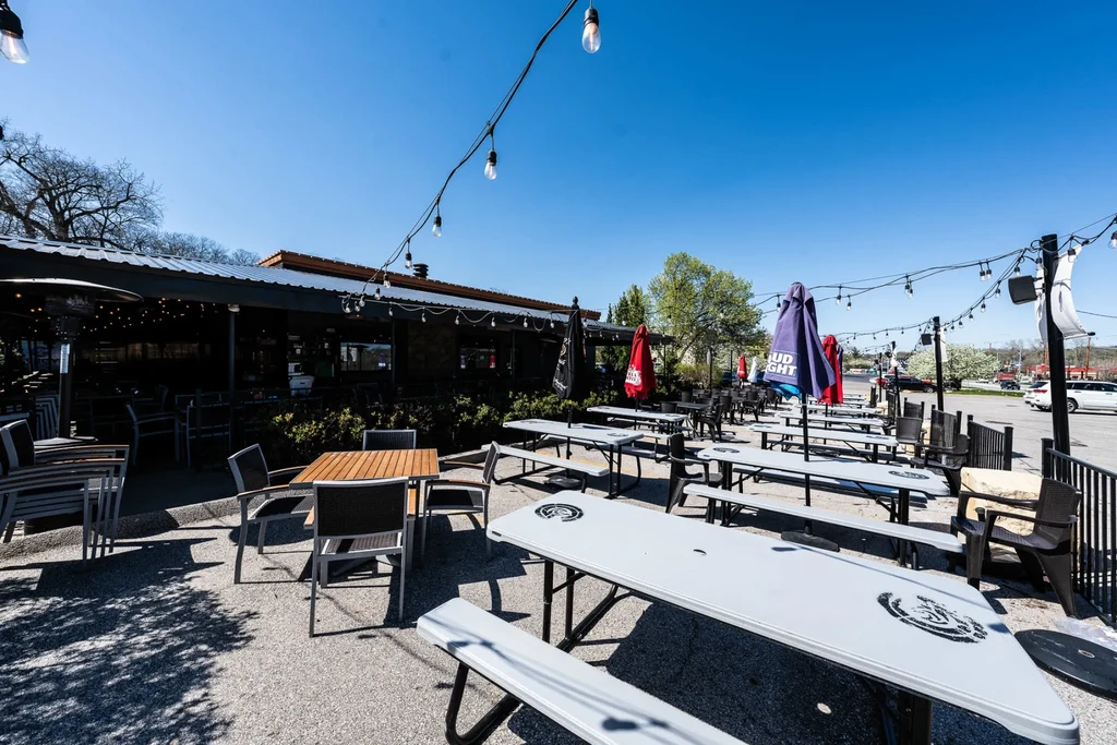 Sunny outdoor restaurant patio with tables, chairs, umbrellas, string lights.
