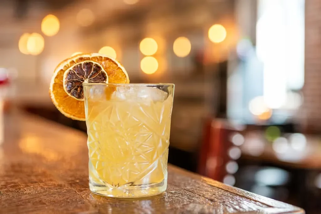 Light yellow cocktail in patterned glass with dried oranges on a wooden bar.