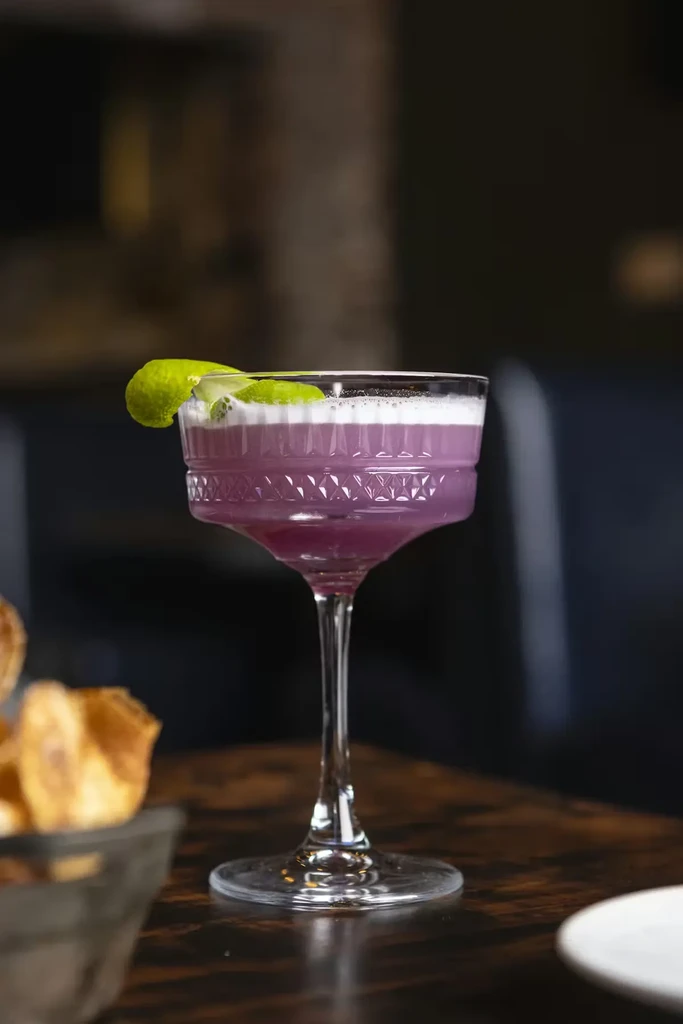 Purple cocktail with white foam and lime peel on a wooden table.