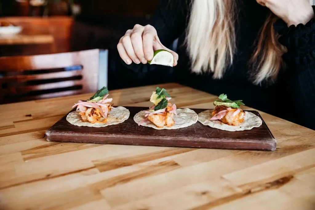 Hands squeeze lime over three grilled shrimp tacos on a wooden board.