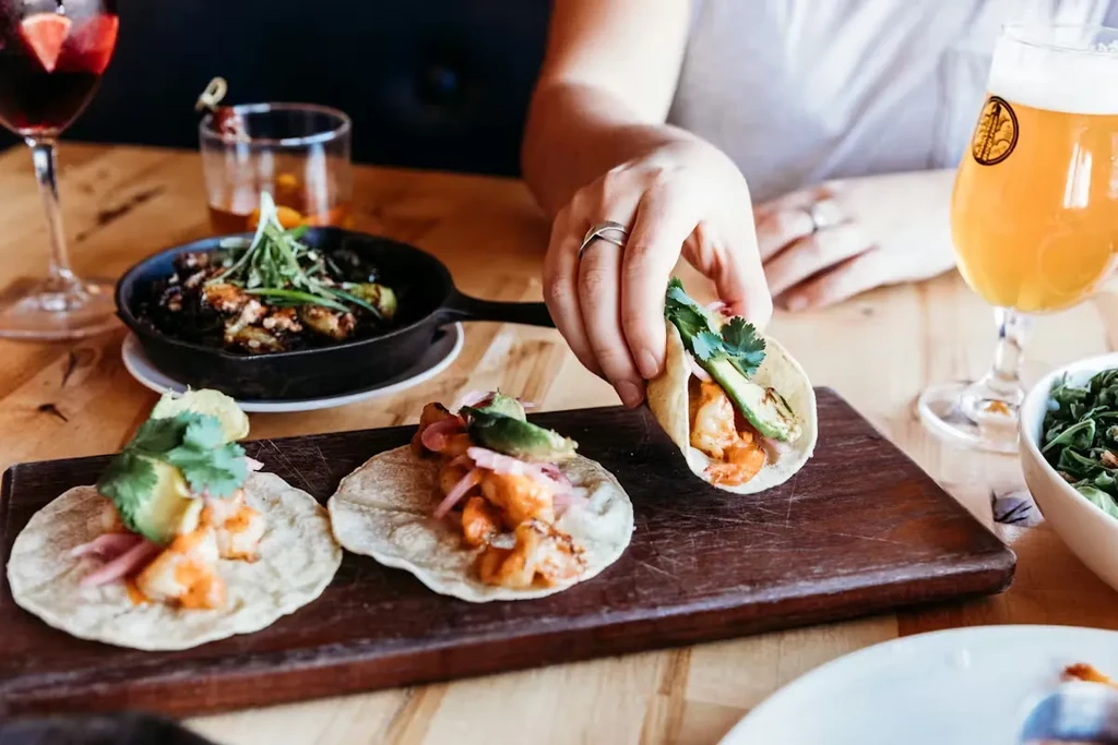 Three shrimp tacos, one held by a hand, with drinks and vegetables.