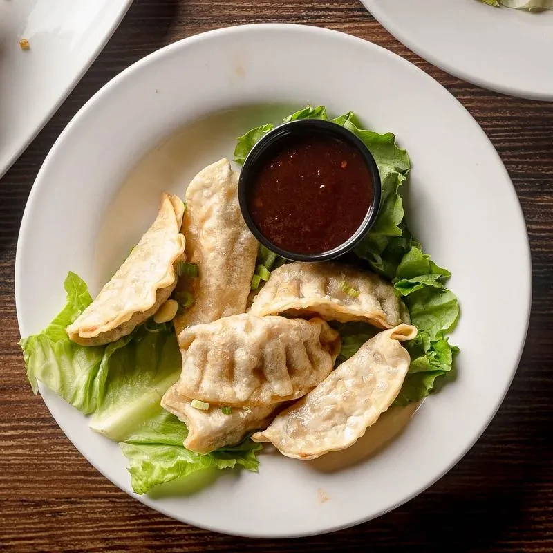 Golden-brown pan-fried dumplings on lettuce with dipping sauce in a bowl.