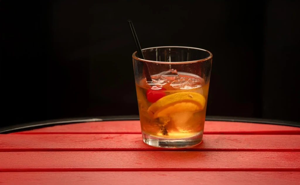 Amber cocktail with ice, maraschino cherry, citrus, straw on red table.