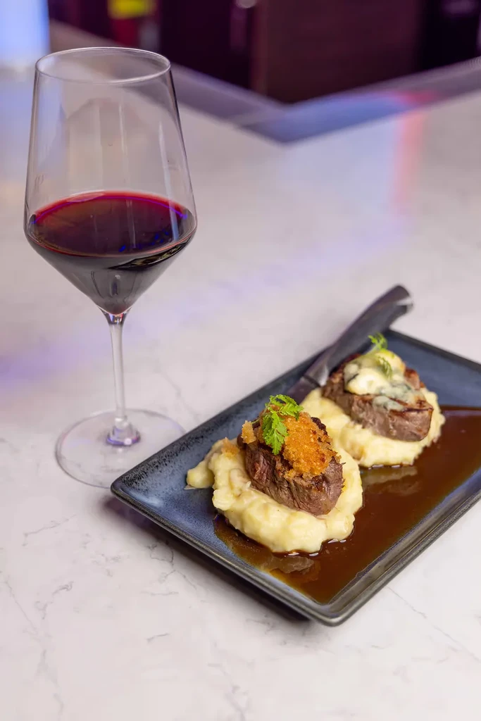 Two garnished steaks, mashed potatoes, and red wine on a dark plate.