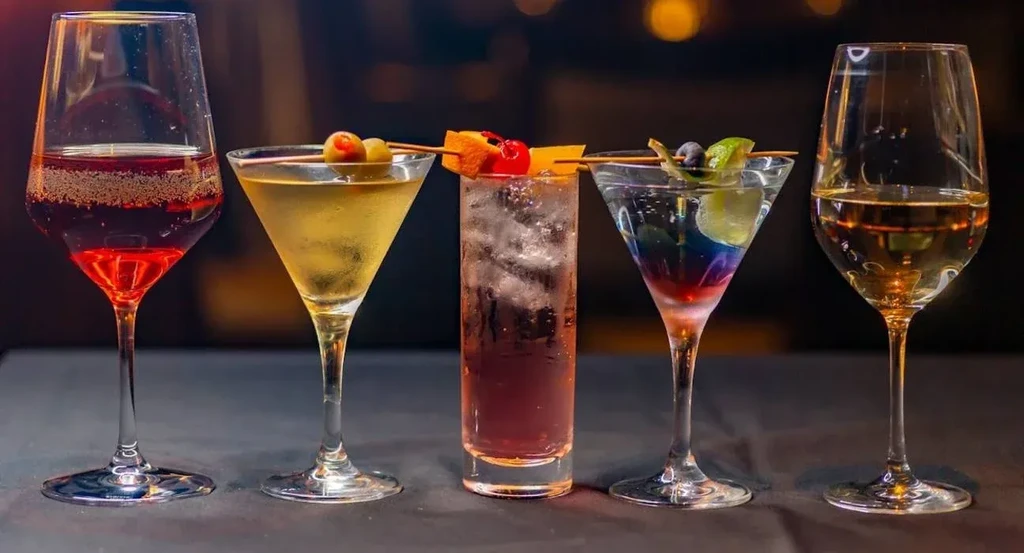 Five diverse cocktails and wines arranged in a line on a dark bar.