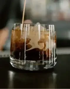 Creamy white liquid pouring into dark iced cocktail in textured glass.