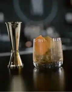 Gold jigger and fluted glass with amber cocktail, ice, orange peel.