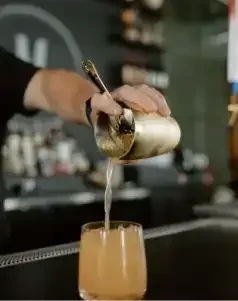 Close-up of golden shaker pouring amber liquid into an ice-filled glass.
