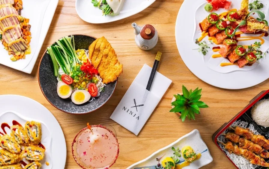 Table full of Asian noodles, sushi, skewers, and a pink cocktail.
