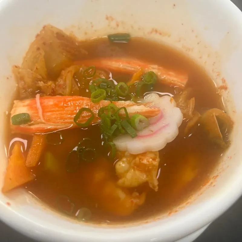 Savory reddish-brown soup with crab sticks, kimchi, fish cake, and green onions.