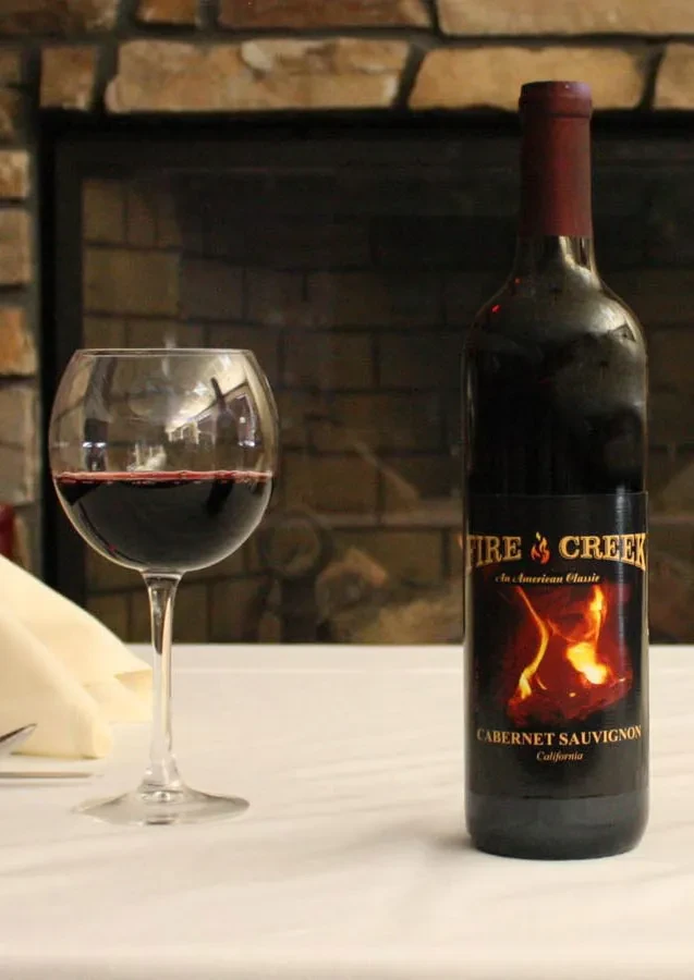 Red wine bottle labeled 'Fire & Creek' and glass by a stone fireplace.