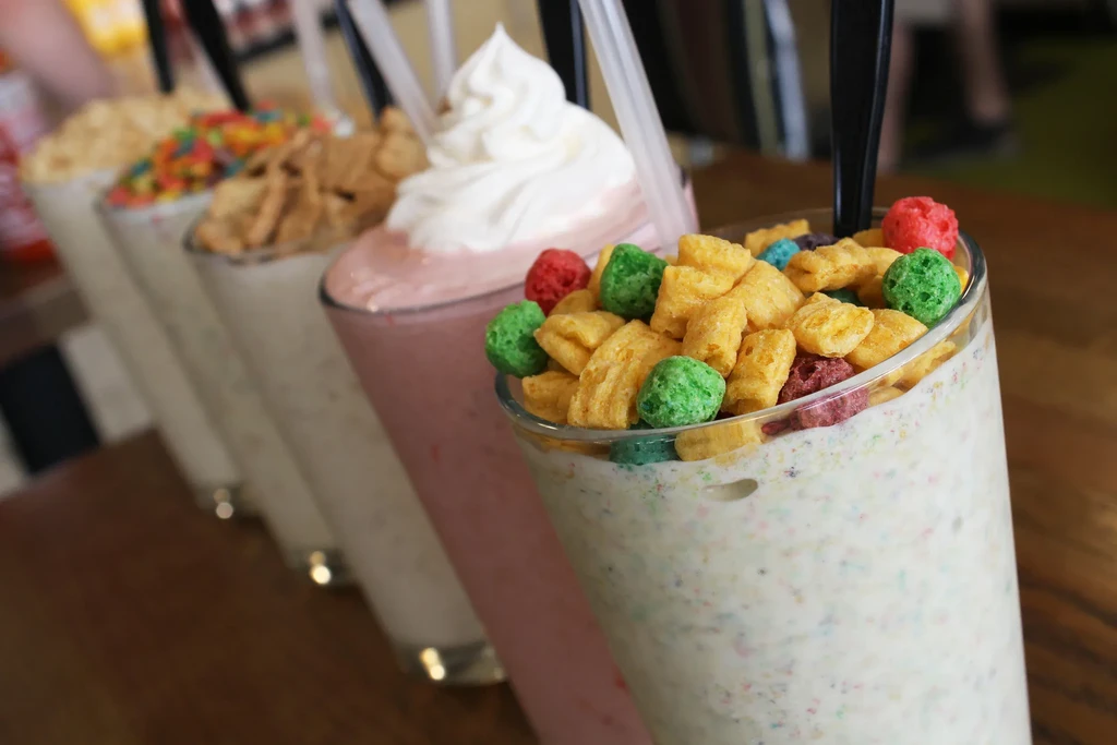 Five colorful milkshakes in clear glasses, topped with various cereals.