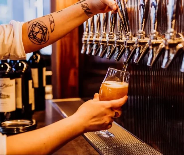Tattooed hands pouring amber beer from a tap into a glass.
