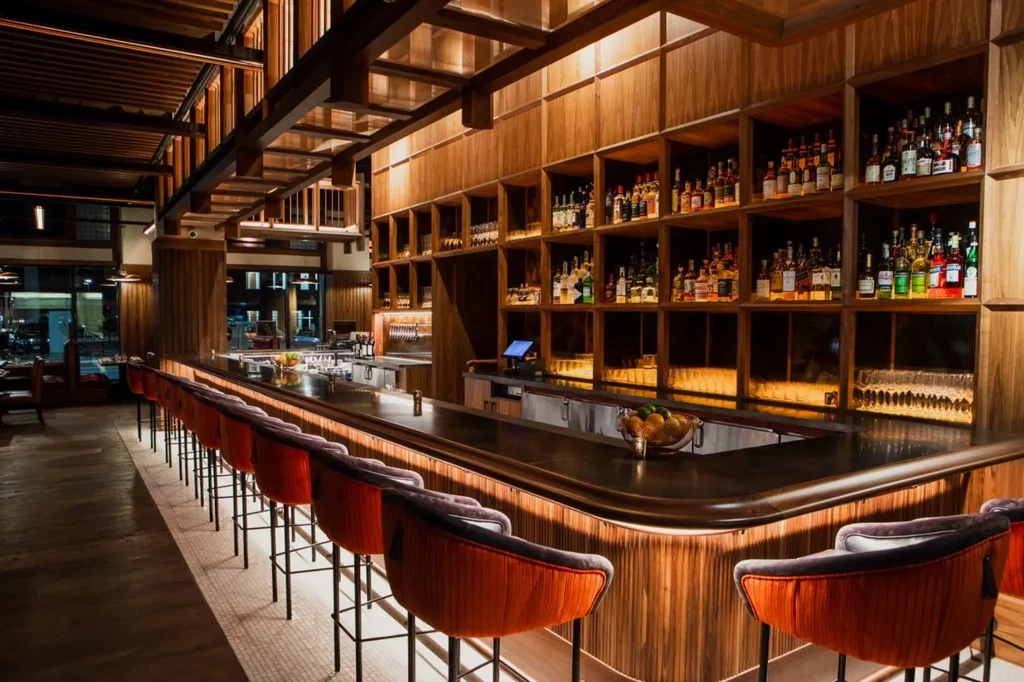 Empty elegant bar: dark counter, orange stools, wood liquor shelves.