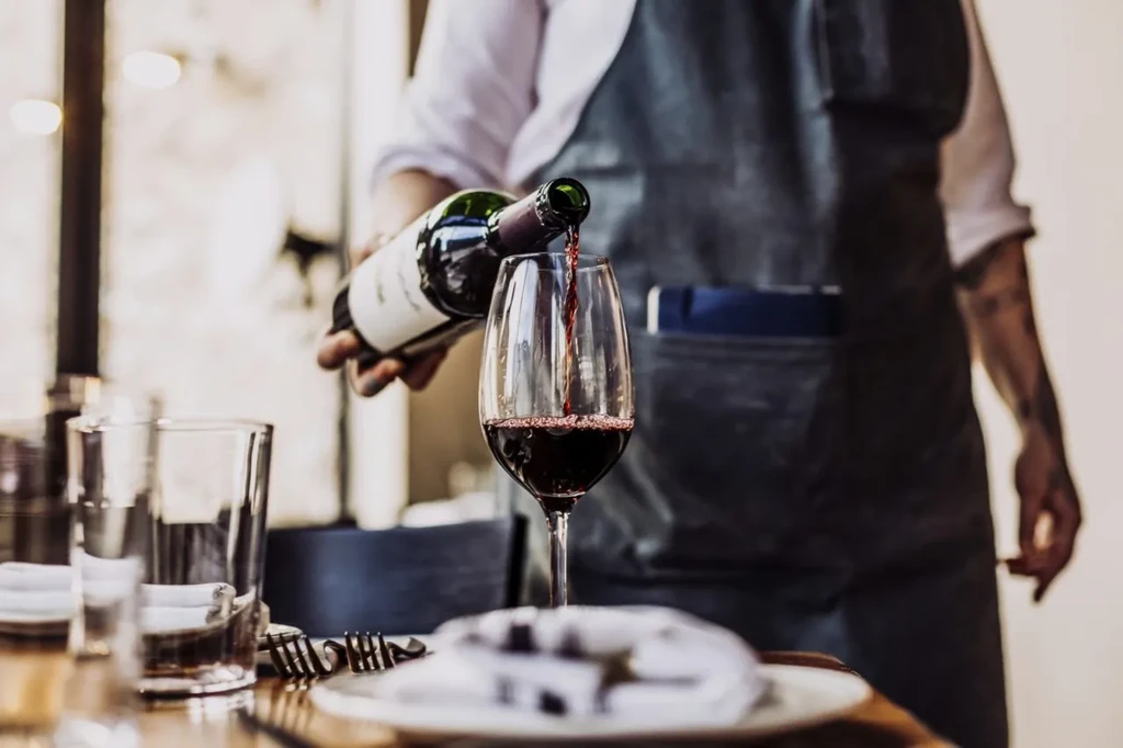 Server pouring red wine from bottle into glass at dining table.