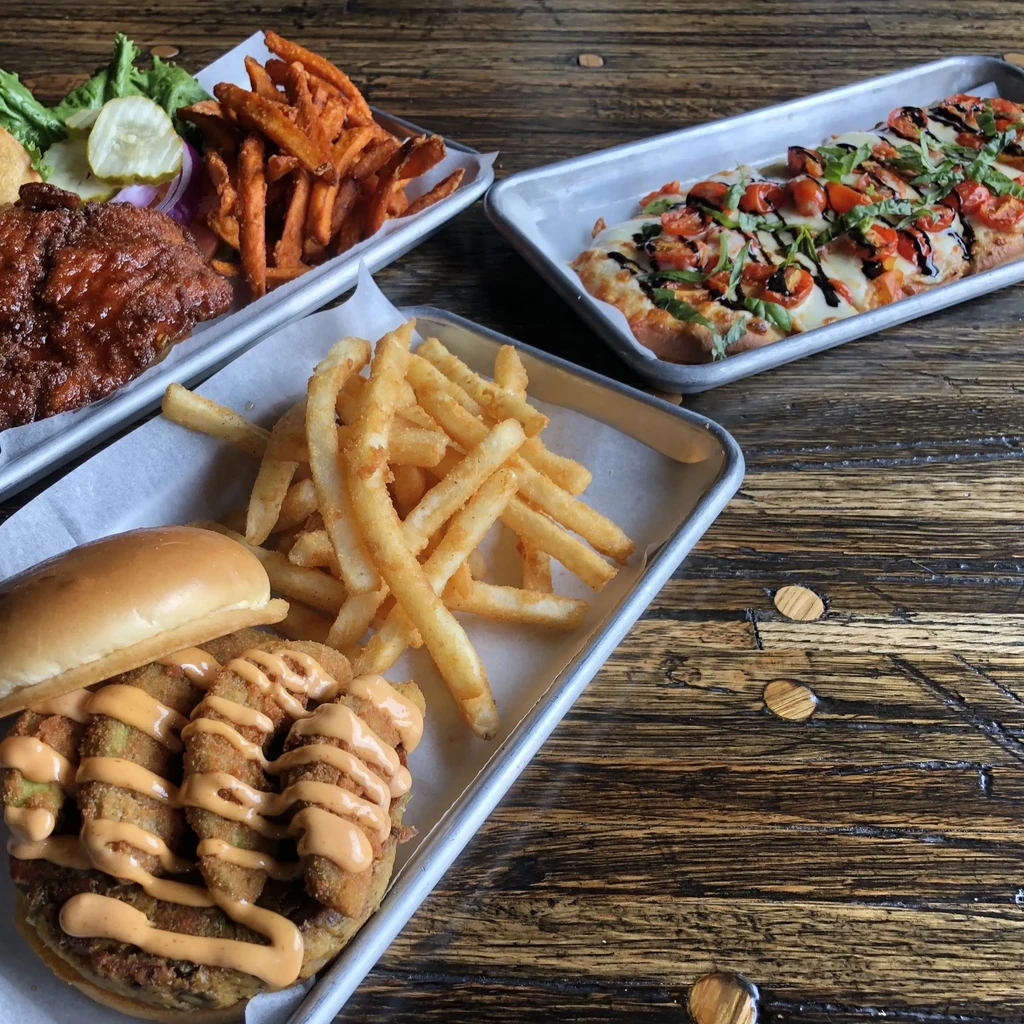 Three metal trays: sandwich, BBQ, flatbread, on a rustic wooden table.