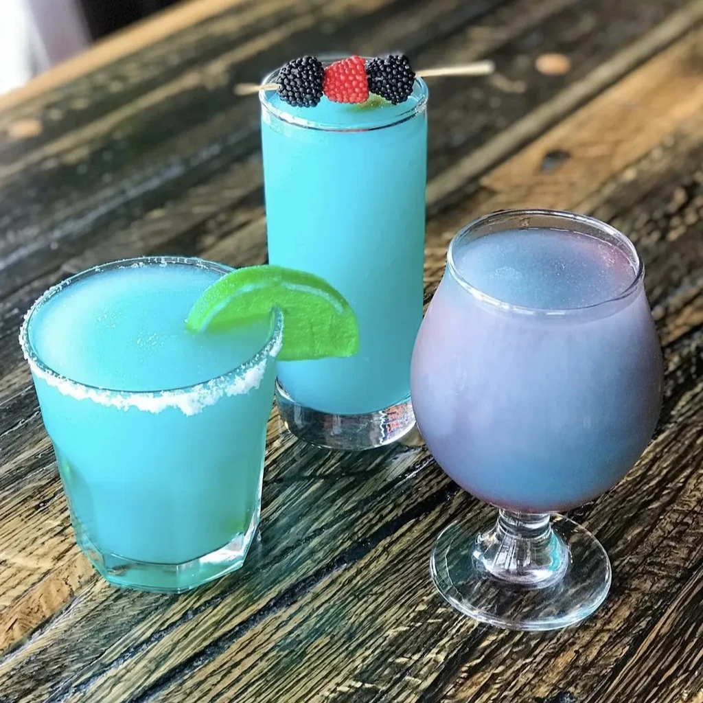 Colorful frozen drinks: two turquoise, one lavender, with garnishes on wood.