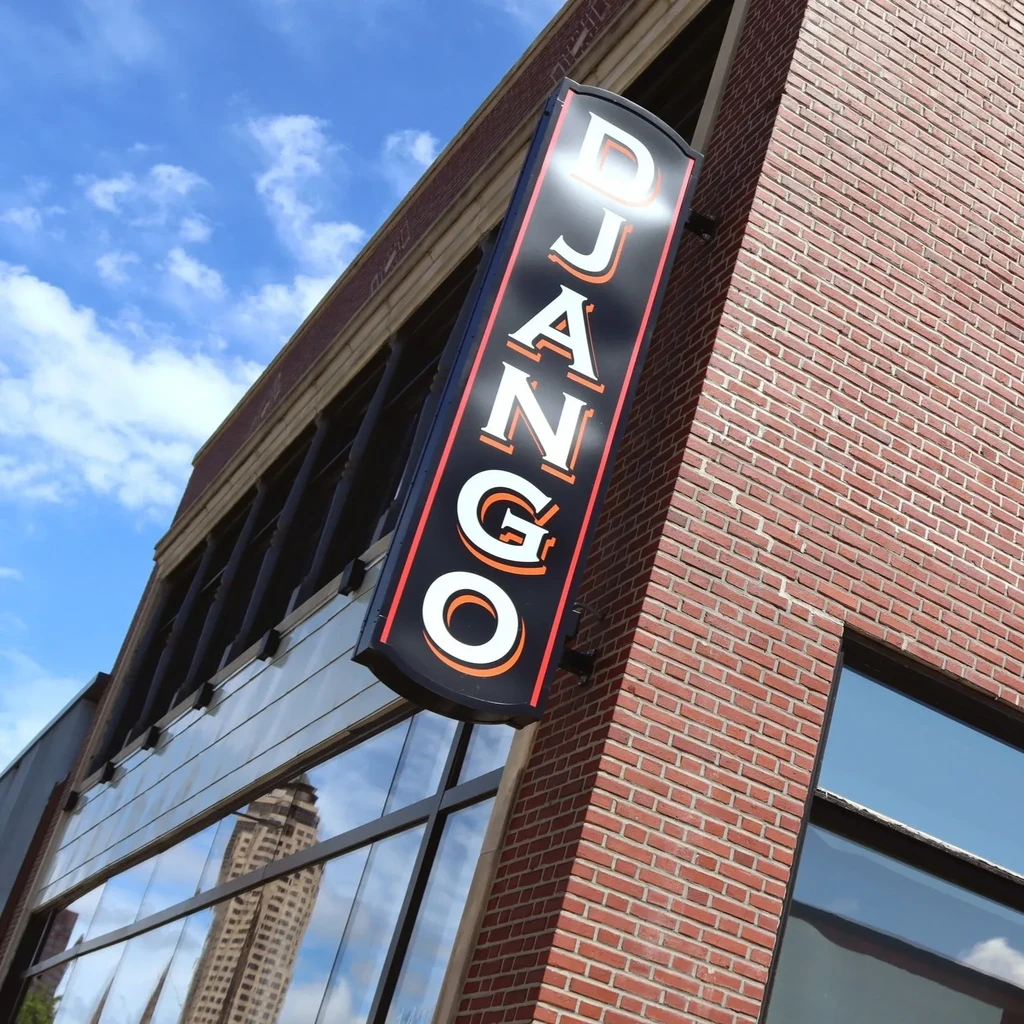 Vertical black "DJANGO" sign on a brick building with window reflections.