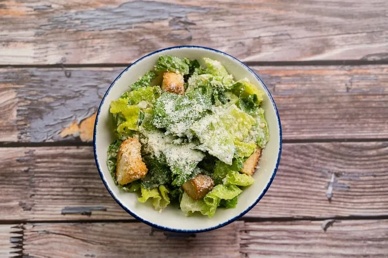 Overhead Caesar salad in a white bowl on a rustic wooden table.