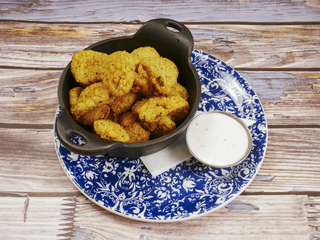Fried pickles/okra in skillet with dipping sauce on a patterned plate.