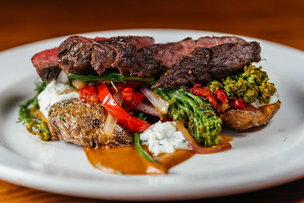 Sliced medium-rare steak, roasted vegetables, and sauces on a white plate.