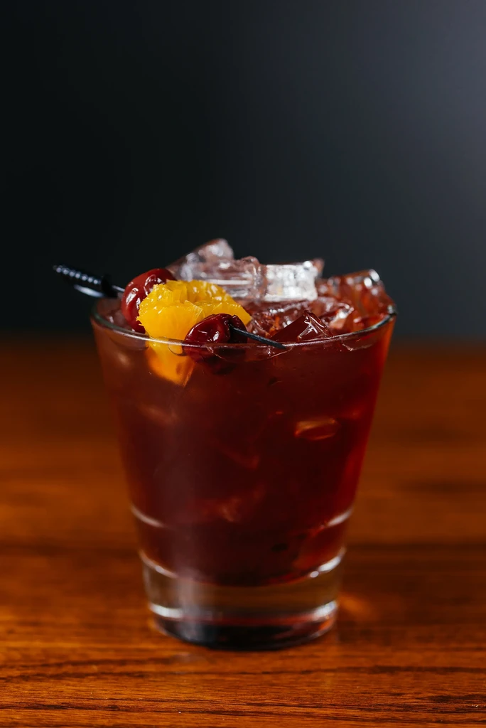 Reddish-brown cocktail with ice, cherries, and orange slice on wood.