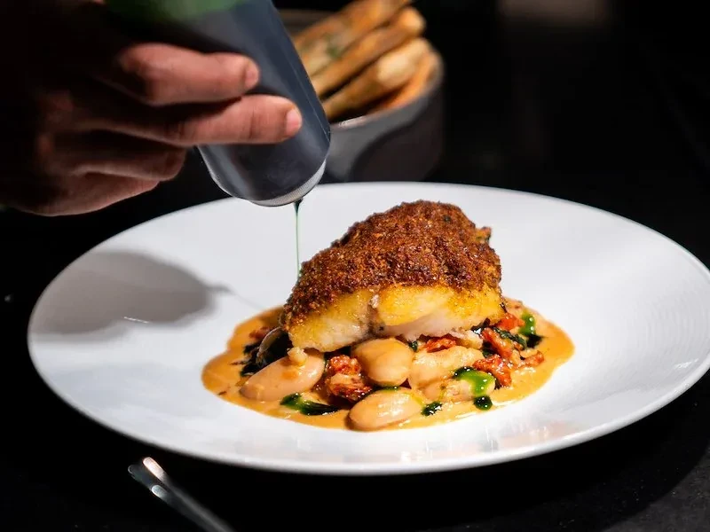 Hand drizzles green sauce onto a plated crispy fish and vegetables.