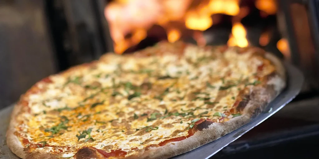 Bubbly cheese pizza on a peel in front of a fiery wood oven.