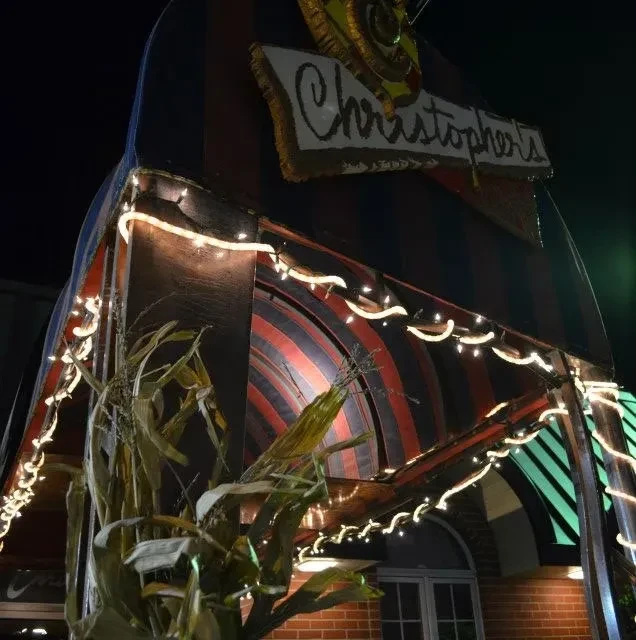 Lit 'Christopher's' restaurant entrance, red-striped awning, string lights, and corn stalks.
