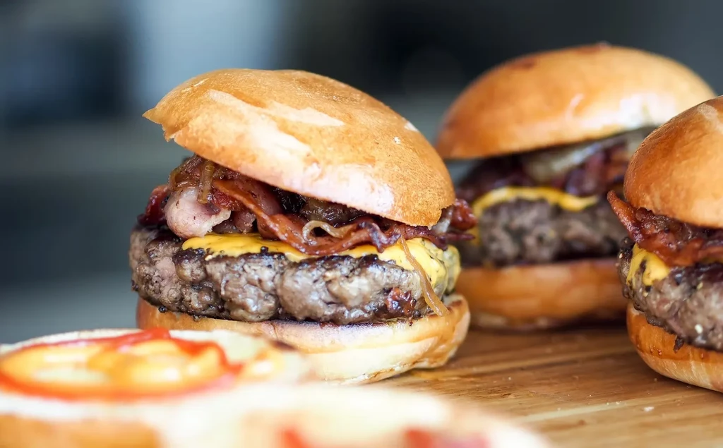 Close-up of multiple beef sliders with cheese, bacon, onions on wooden board.