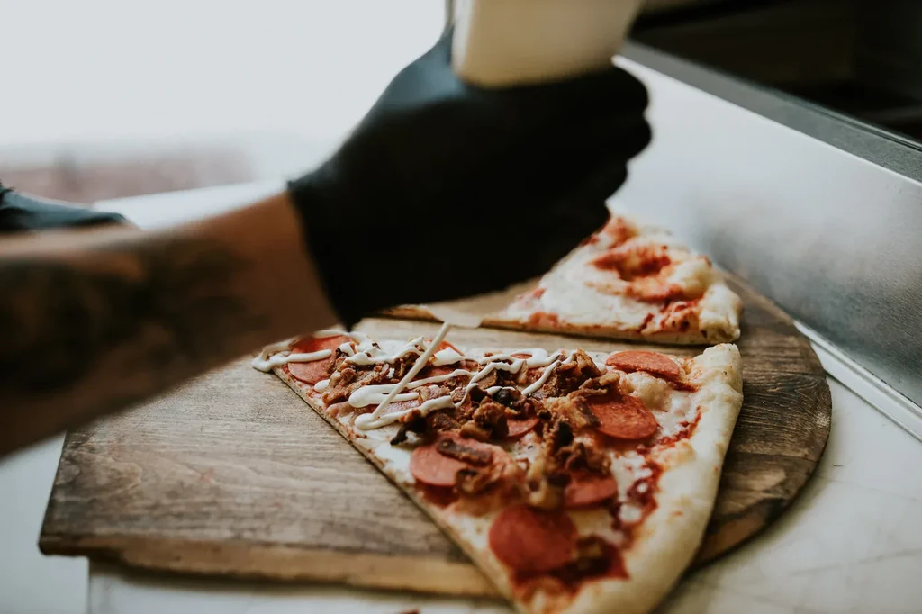 Gloved hand drizzles sauce on pepperoni and shredded meat pizza slice.