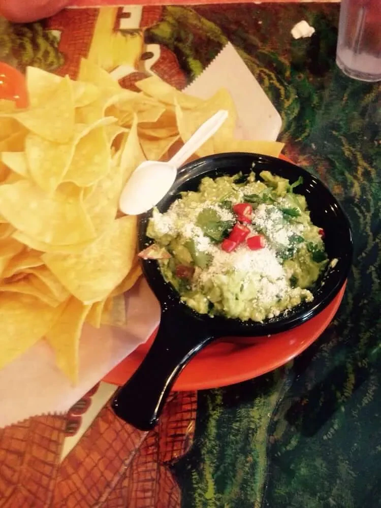 Top-down view of guacamole, chips, cheese, chilies on patterned table.