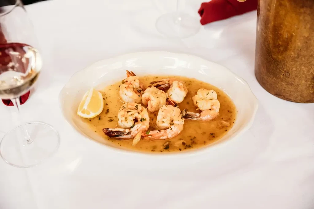 Dish of cooked shrimp in sauce with lemon, wine glass.