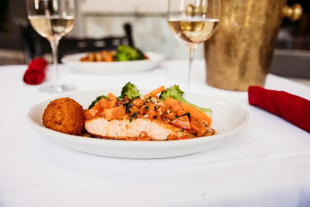 Plated salmon with colorful vegetables, broccoli, carrots, and a croquette.