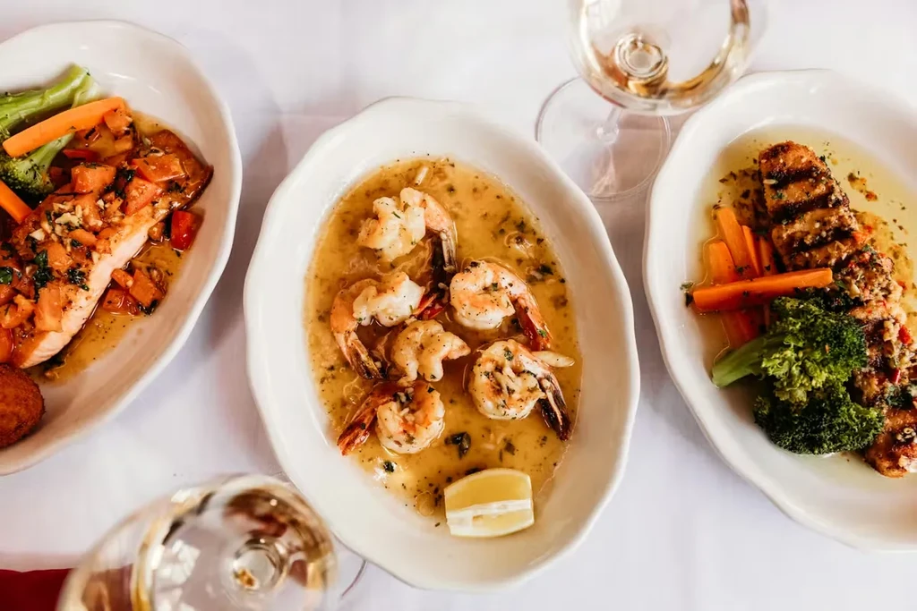 Restaurant table with shrimp, salmon, vegetables, and two glasses of wine.