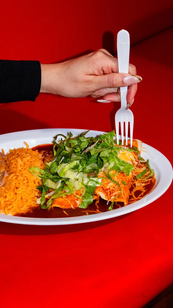 Enchiladas, rice, beans on a plate; hand holds fork, red background.
